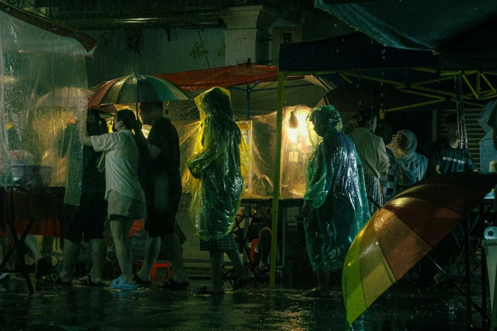 Shoppers in rain ponchos at a Malaysian night market during heavy rainfall, illustrating the constant moisture that demands high-performance waterproofing